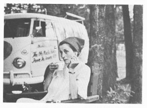 Photo of Margarete sipping tea during a roadside stop. Bill's Volkswagen bus is parked behind in a wooded area.